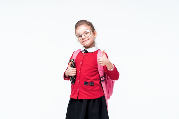 Entzückendes kleines Mädchen in der roten Schuljacke, im schwarzen Kleid, in der abgerundeten Brille und im Rucksack, der Daumen oben zeigt
