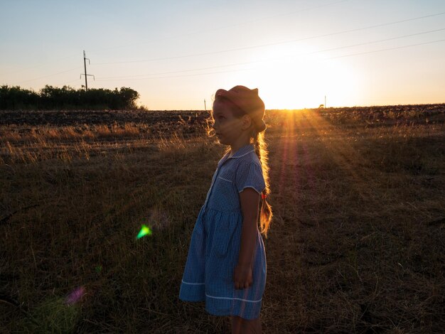 Entzückendes kleines Mädchen in einem blau karierten Sommerkleid mit Strohhut auf einer Wiese auf dem Land. Kind mit langem blonden Zopf