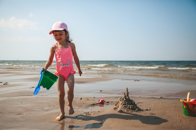 Entzückendes kleines Mädchen in einem rosafarbenen Badeanzug, der mit Sand am Strand einer Ostsee spielt