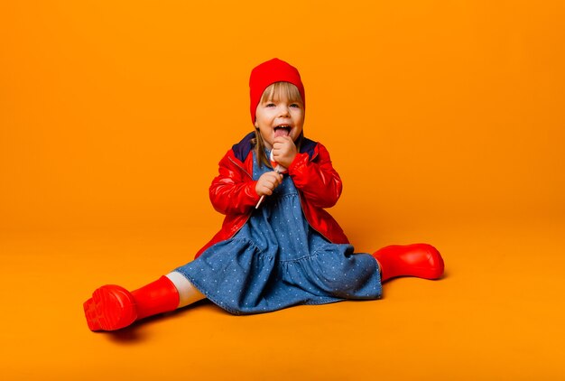 Entzückendes kleines Mädchen in einer roten Jacke und Stiefeln, die einen Lutscher auf gelbem Hintergrund halten. Herbst Konzept.