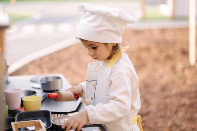 Entzückendes kleines Mädchen in Kochmantel und Mütze kocht in der Kinderspielzeugküche, die auf kleinen Kindern spielt