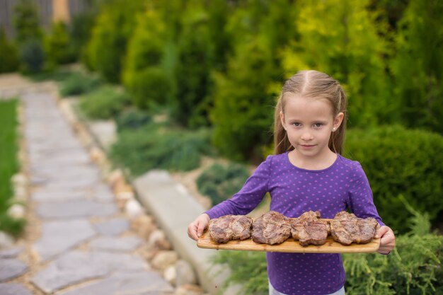 Entzückendes kleines Mädchen mit gegrillten Steaks in den Händen im Freien