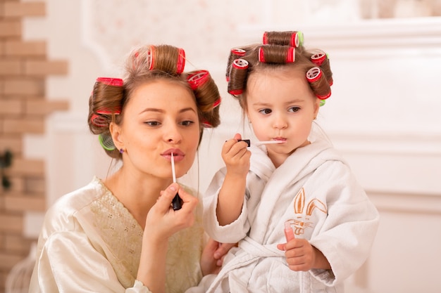 Entzückendes kleines mädchen mit ihrer mutter in lockenwicklern tragen make-up auf. mutter bringt tochter bei, kosmetik zu benutzen. schönheitstag. mädchen sind solche mädchen.