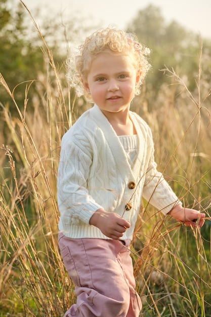 Entzückendes kleines Mädchen mit lockigem Haar und weißem Pullover, das auf einem grünen Rasen in der sonnigen Sommerlandschaft steht und in die Kamera blickt