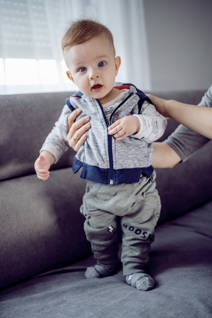 Entzückendes Kleinkind, das mit Hilfe seiner Mutter auf der Couch steht.