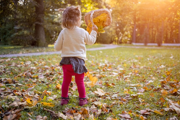 Entzückendes Kleinkindmädchen, das an einem sonnigen Tag mit gelben Ahornblättern im Herbstpark spielt