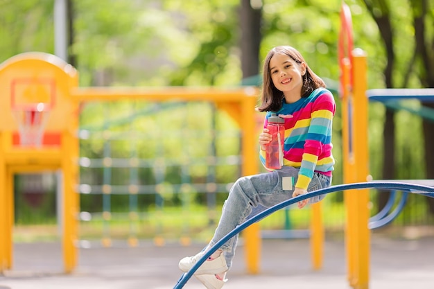 Entzückendes lächelndes kleines Mädchen 56 Jahre alt, das mit einer Plastikflasche Wasser auf dem Spielplatz sitzt