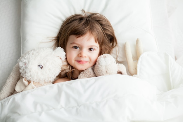 Entzückendes lächelndes kleines Mädchen, das im weißen Bett mit Kaninchenspielzeug liegt