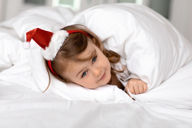 Entzückendes lustiges kleines Mädchen mit blauen Augen in rotem Weihnachtsmann-Hut liegt im Bett unter einer weißen flauschigen Decke