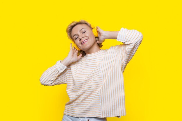 Entzückendes Nahaufnahmefoto einer blonden Dame, die das Nachdenken und Genießen der Zeit auf einer gelben Studiowand hört