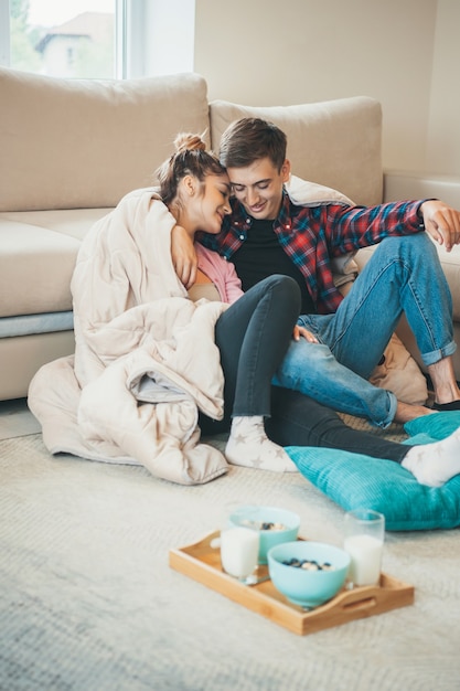 Entzückendes Paar umarmt sich auf dem Boden in der Nähe der Couch, bevor es Müsli mit Milch isst