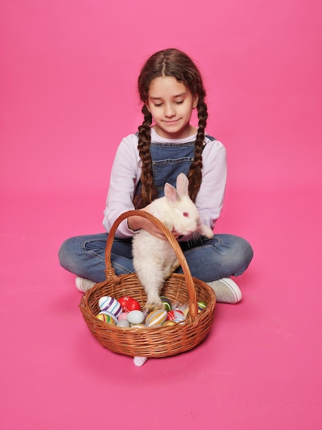 Entzückendes Schulmädchen spielt mit süßem Hasen und legt sie in einen Korb, Posen im Studio auf rosafarbenem Hintergrund