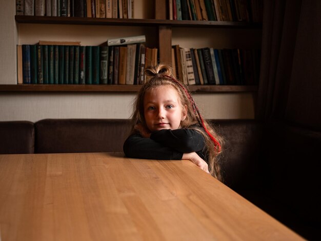 Entzückendes, stilvolles kleines Mädchen mit langen, bunten Haaren im Hintergrund eines Bücherregals im modernen Bibliothekscafé. Junger Leser sitzt allein auf einer bequemen Couch am Tisch. Authentisches, fröhliches, offenes Kinderporträt