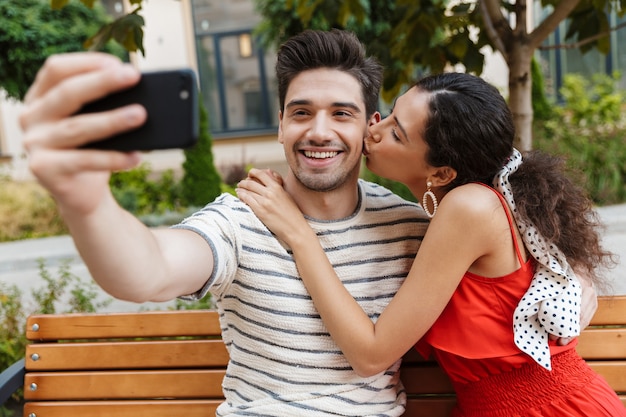 Entzücktes junges Paar, das ein Selfie-Foto auf dem Handy macht und sich küsst, während es auf einer Bank in der grünen Straße sitzt?