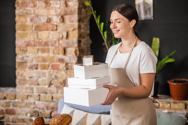 Envases, productos de panadería. Mujer alegre bastante ordenada que lleva cajas de embalaje de panadería en la acogedora habitación