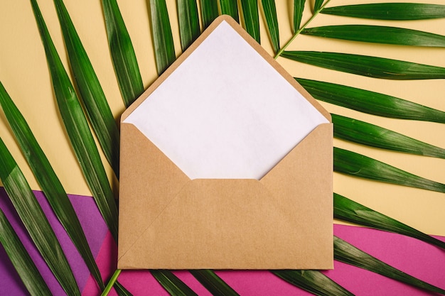 Envelope de papel kraft marrom com branco cartão vazio em folhas de palmeira, rosa, roxo e creme fundo amarelo, carta em branco maquete