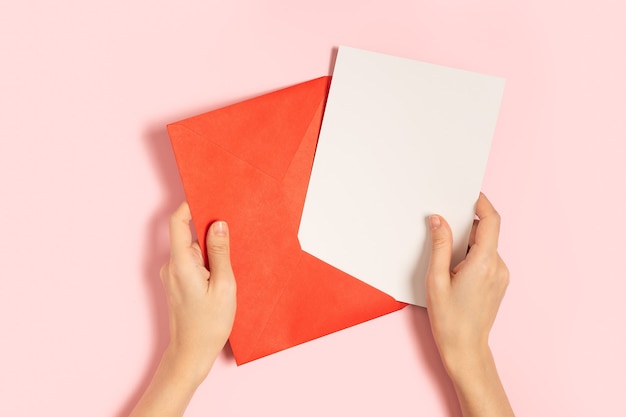 Envelope de papel vermelho com maquete de nota branca em branco nas mãos da mulher, dentro em fundo rosa pastel