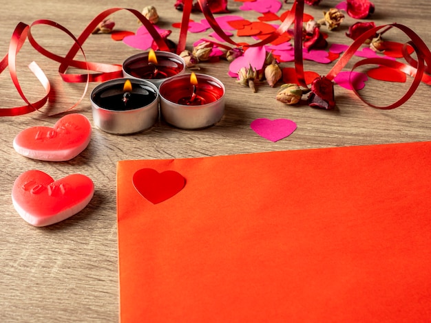 Envelope vermelho ao lado de velas com pétalas de rosa vermelhas e corações na mesa