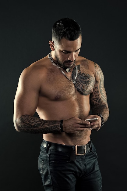 Enviar mensagem Homem bonito em forma enviar mensagem smartphone Atleta tatuado musculoso parece atraente Conceito de comunicação de mensagens Homem bonito musculoso sem camisa com jeans sobre fundo escuro