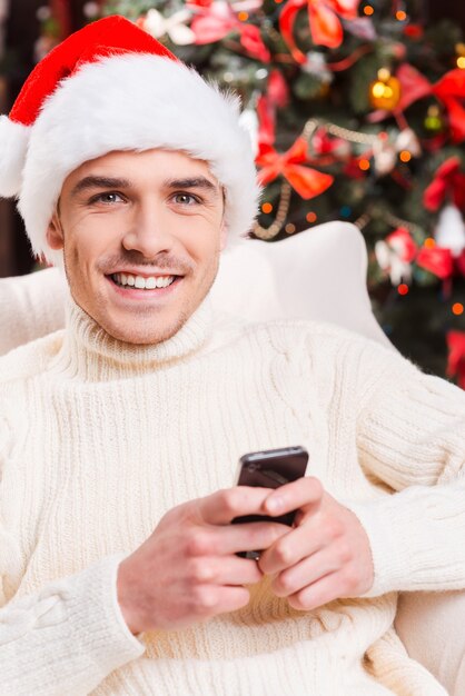 Enviar mensajes de texto a su más cercano. Apuesto joven con sombrero de Santa enviando mensajes de texto en su teléfono móvil y sonriendo con árbol de Navidad en el fondo