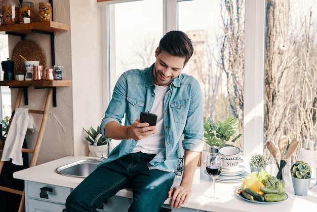 Enviar mensajes de texto a su novia. Apuesto joven en ropa casual sonriendo y usando su teléfono inteligente mientras está sentado en la cocina en casa