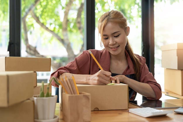 Envio de compras on-line pequeno empresário de inicialização escrevendo endereço em caixa de papelão no local de trabalho Mulher asiática freelance pequena empreendedora PME trabalhando com caixa em casa