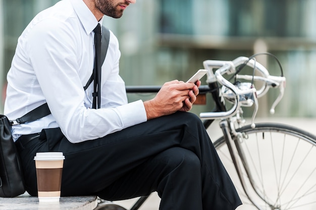 Envío de mensaje comercial. Imagen recortada del joven empresario sosteniendo un teléfono móvil mientras está sentado cerca de su bicicleta al aire libre