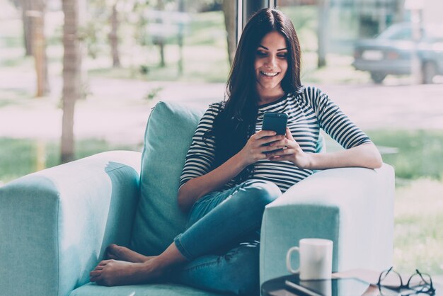 Envío de mensaje encantador. Hermosa joven sonriente mirando su teléfono inteligente mientras está sentado en una silla grande y cómoda en casa