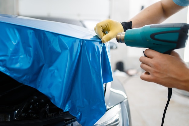 Envoltorio de coche, hombre con secador instala lámina protectora de vinilo o película en primer plano del capó del vehículo. Trabajador hace detalles automáticos