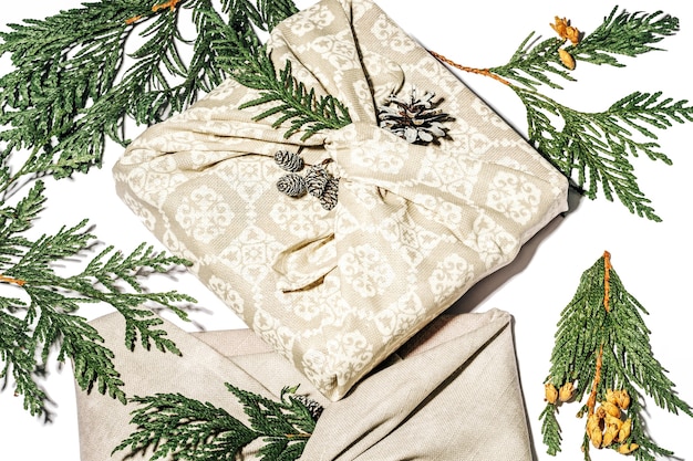 Envoltorio de regalo sin desperdicio al estilo tradicional japonés furoshiki. Paquete de regalo beige hecho a mano para Navidad con tela, thuja y cono en blanco. Concepto ecológico decoración ecológica Navidad. De cerca.