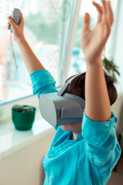 Envolvido na escola levantando as mãos no ar jogando um jogo usando grandes óculos VR em pé perto da janela
