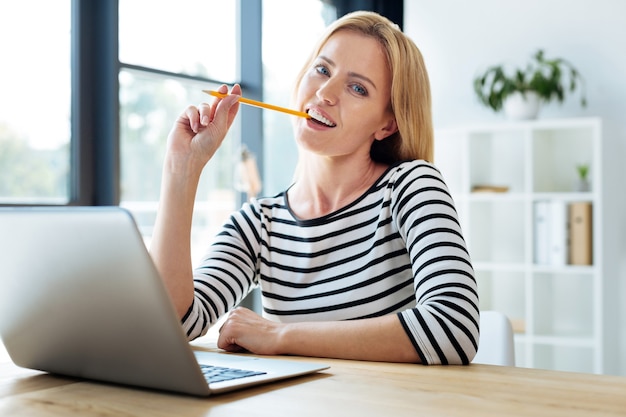 Envolvido no trabalho. Mulher inteligente e simpática sentada à mesa e olhando para você enquanto morde o lápis