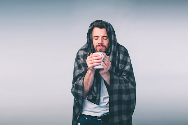 Envuelto en la cálida manta o cuadros, enfermo, joven mocoso con gafas bebiendo té curativo caliente