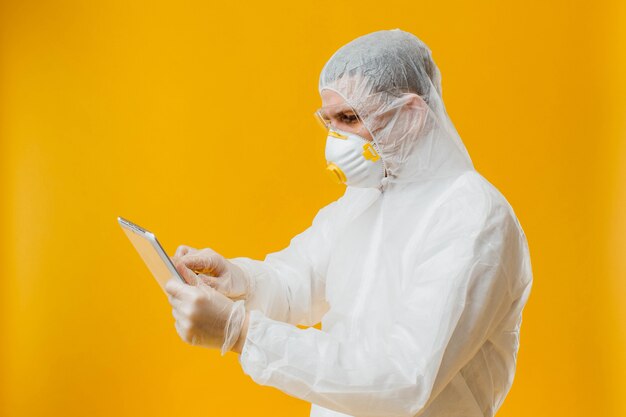 Epidemiólogo en traje de materiales peligrosos y máscara de respiración con tableta digital en pared amarilla