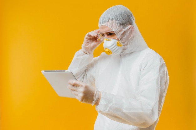 Epidemiólogo en traje de materiales peligrosos y máscara de respiración con tableta digital en pared amarilla