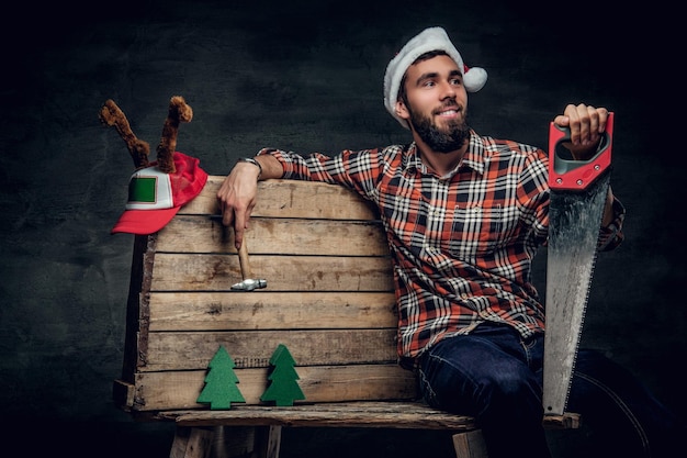 Época de natal. retrato de estúdio de macho barbudo com chapéu de papai noel segura o serrote e senta-se em uma paleta com abetos.