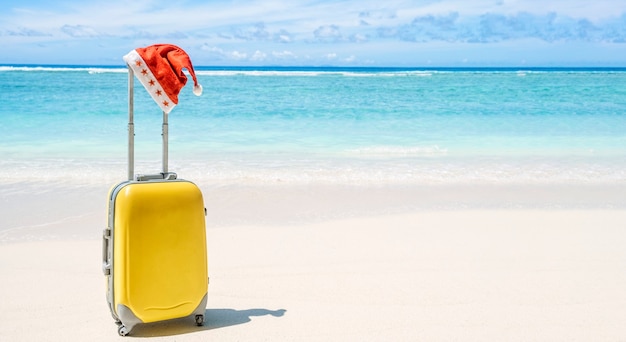 Equipaje de mano con ruedas rígido amarillo con gorro de Papá Noel rojo en una playa de arena