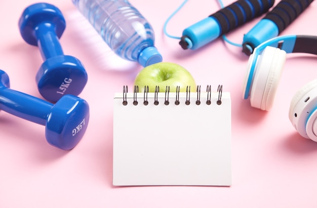 Equipamentos de ginástica. Bloco de notas, maçã, fones de ouvido, garrafa de água rosa