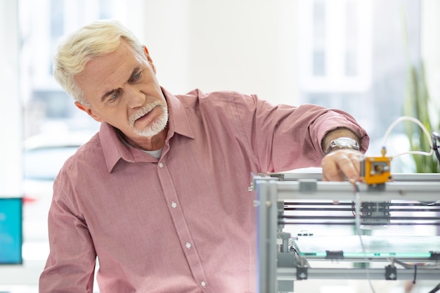 Equipamentos de última geração. homem idoso encantador usando uma impressora 3d no escritório e controlando seu trabalho