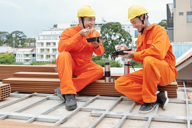 Equipe de construtores sorridentes almoçando e bebendo café no telhado após uma manhã produtiva em con ...