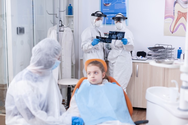 Equipe de dentista, olhando para a radiografia dentária do paciente, vestida com macacão durante o surto de coronavírus. Estomatologista durante covid19 vestindo terno ppe fazendo procedimento dentário de criança sentada