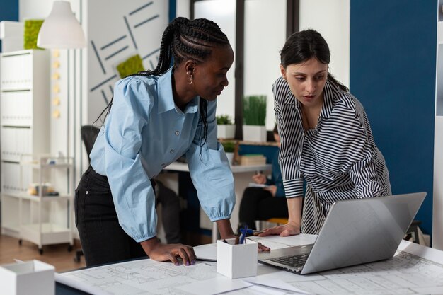Equipe de duas arquitetas profissionais trabalhando no computador laptop com planos de construção na mesa em um escritório de arquitetura. Engenheiros projetando plantas para projeto imobiliário.