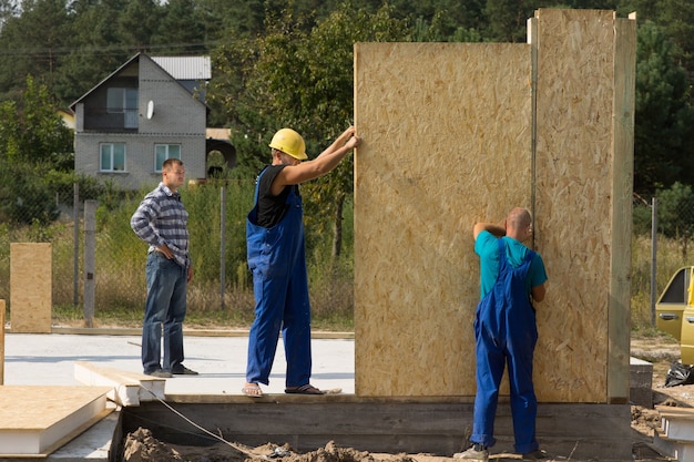 Equipe de jovens construtores erguendo painéis de parede pré-fabricados no canteiro de obras de uma nova casa