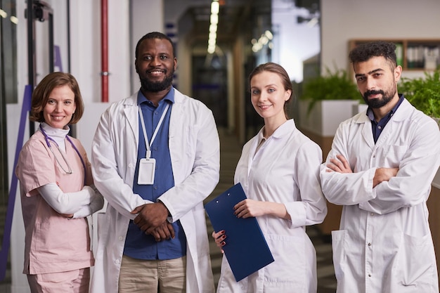 Foto equipe de jovens trabalhadores de saúde interculturais felizes de uniforme olhando para a câmera enquanto estão em fila no corredor de clínicas modernas