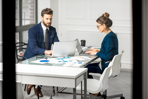 Equipe de marketing ou analista vestida com ternos, trabalhando com gráficos de papel e laptops no interior do escritório branco