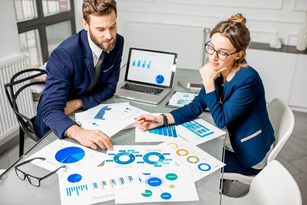 Equipe de marketing ou analista vestida com ternos, trabalhando com gráficos de papel e laptops no interior do escritório branco