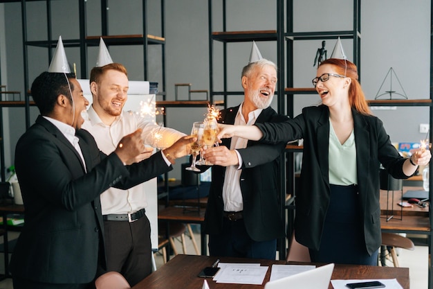 Equipe de negócios alegre de colegas de trabalho multiétnicas segurando luzes de Bengala tilintando copos de champanhe