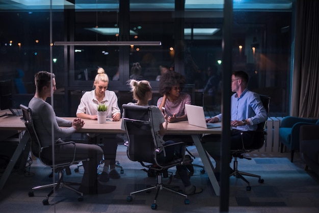 Equipe de negócios de inicialização multiétnica em reunião no interior do escritório noturno moderno brainstorming, trabalhando no laptop