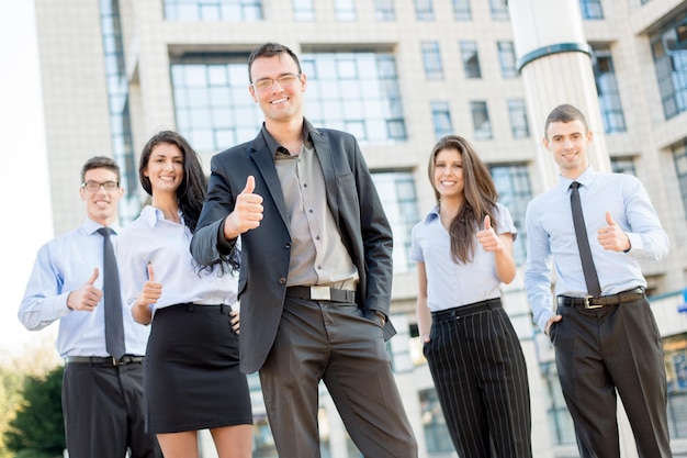 Equipe de negócios jovem, elegantemente vestida do lado de fora na frente do prédio com os polegares para cima. Olhando para a câmera.