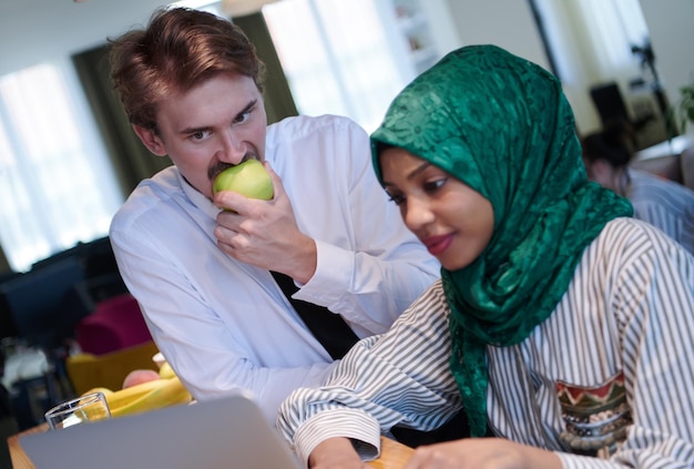 equipe de negócios multiculturais internacionais homem comendo maçã mulher muçulmana africana vestindo hijab verde bebendo chá enquanto trabalhava juntos no computador portátil na área de relaxamento no plano aberto moderno startu